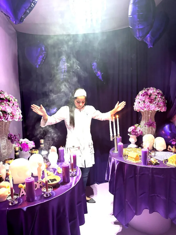 Mãe Jassiara de braços abertos entre duas mesas roxas, com fumaça de defumação e velas